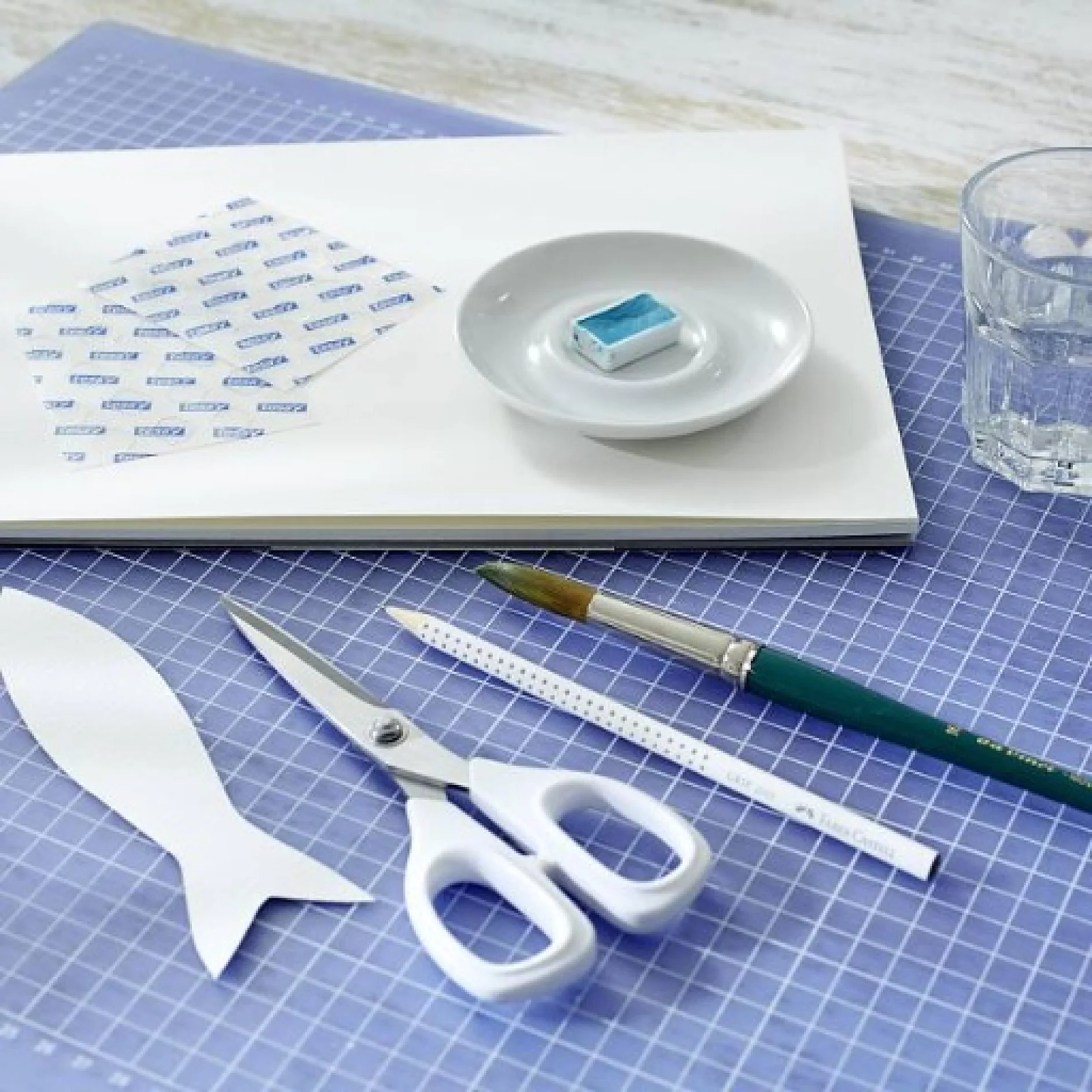 tesa_trendpapier_15_Idee_1_Fische_an_Wand_Schritt_1_1x1 Ein Arbeitsplatz mit Schneidematte, Notizbuch, Lineal, Schere, Bleistift, Pinsel und Radiergummi. Daneben steht ein Glas Wasser. Die Gegenstände sind mit Tesa-Klebeband ordentlich auf der gitterförmigen Oberfläche angeordnet. (This text has been generated by AI)
