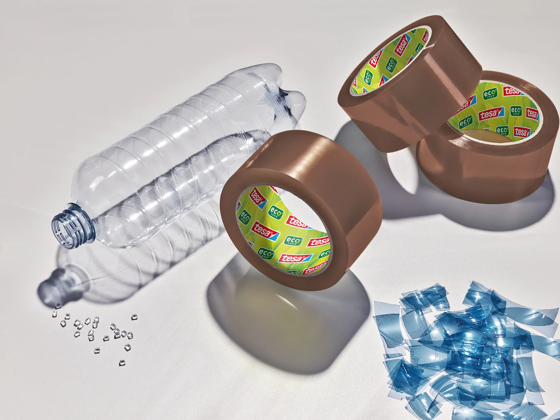 Eine leere Plastikflasche auf der Seite neben kleinen durchsichtigen Plastikperlen, vier Rollen braunem Tesa-Verpackungsband und einem Stapel zugeschnittener Plastikstücke auf einer weißen Oberfläche, die Schatten werfen. (This text has been generated by AI)