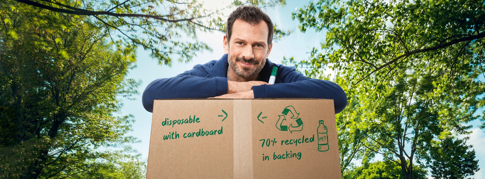 Ein Mann mit dunklem Haar und Bart lehnt an einem großen Karton mit grünem Text: „Einwegkarton mit Karton“ und „70 %+ Recyclingmaterial im Trägermaterial“. Im Hintergrund sind Bäume und ein strahlend blauer Himmel zu sehen. (This text has been generated by AI)