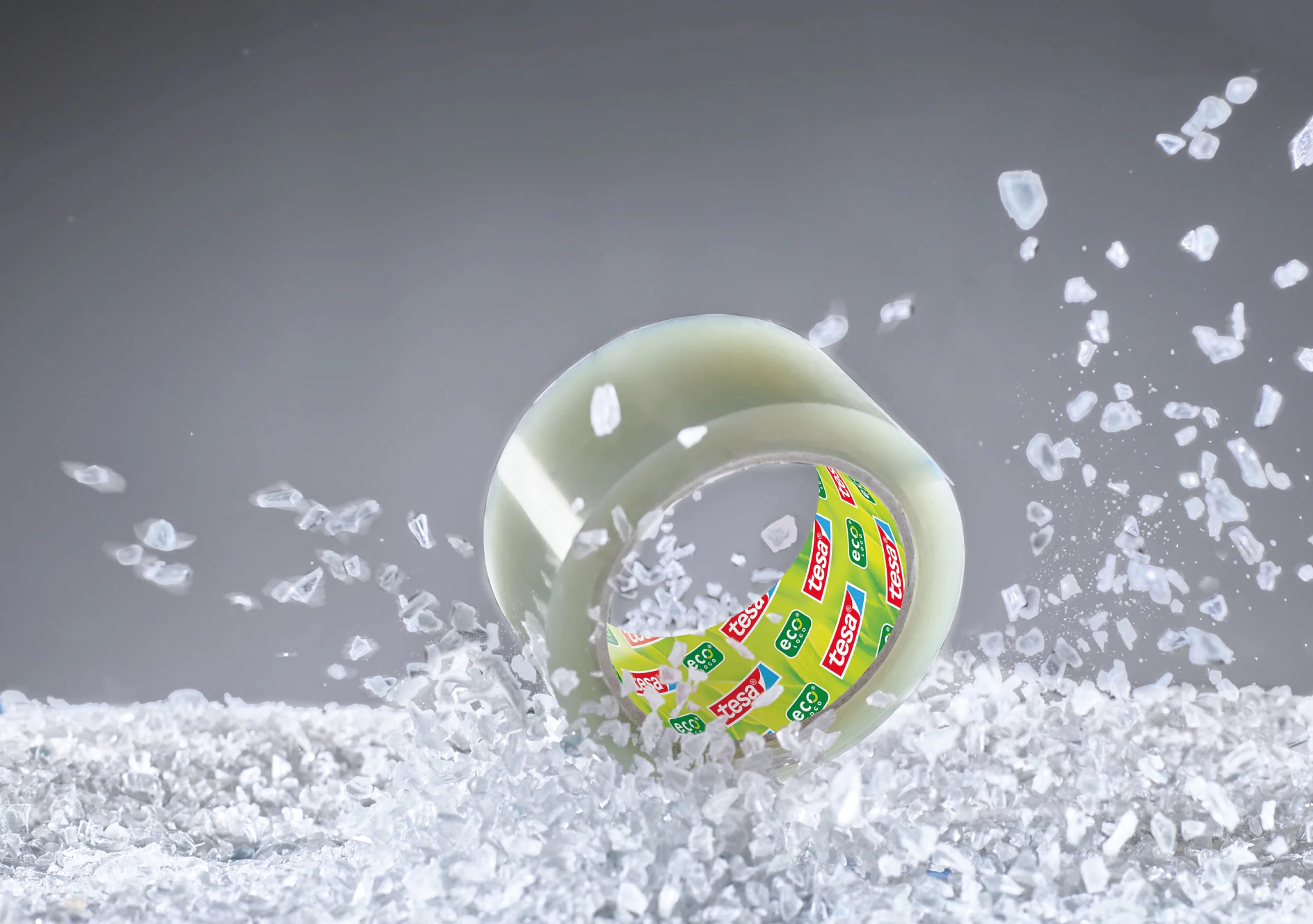 Eine Rolle transparentes Tesa-Klebeband mit farbigem Logo auf der Innenseite ist zwischen verstreuten Eiswürfeln vor einem grauen Hintergrund zu sehen. Das Bild fängt die Bewegung des Eises ein, als ob es gestört worden wäre. (This text has been generated by AI)