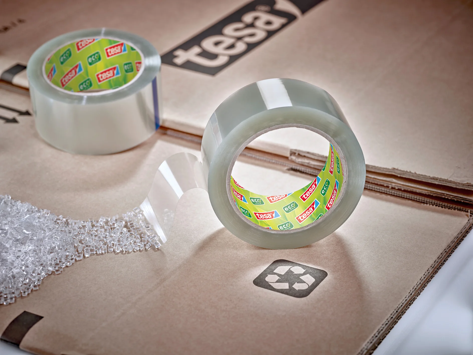 Eine Rolle transparentes Tesa-Klebeband, teilweise abgespult, liegt auf einer Kartonoberfläche. Eine weitere Rolle ist im Hintergrund zu sehen. Auf dem Karton ist ein Recycling-Symbol zu sehen. Das Tesa-Band hat ein Marken-Kernetikett. Auf der linken Seite ist teilweise Luftpolsterfolie zu sehen. (This text has been generated by AI)