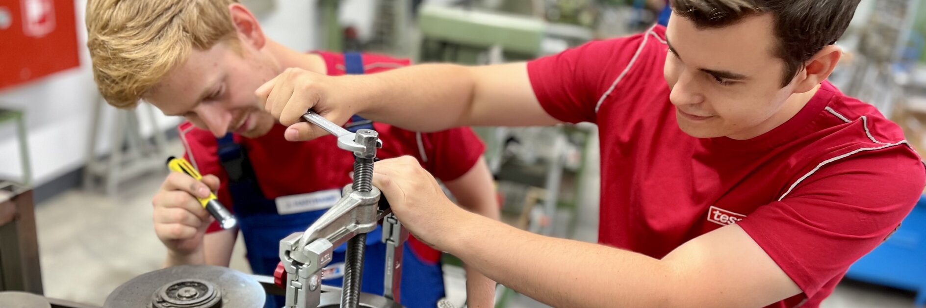 Two young men work together in a workshop. One adjusts a metal part with a wrench, while the other writes with a marker. They are surrounded by machinery and tesa tape. Both wear red shirts and are focused on their task. (This text has been generated by AI)