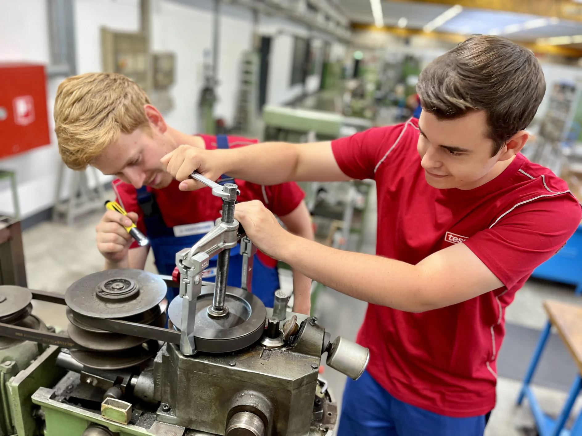 Two young men work together in a workshop. One adjusts a metal part with a wrench, while the other writes with a marker. They are surrounded by machinery and tesa tape. Both wear red shirts and are focused on their task. (This text has been generated by AI)