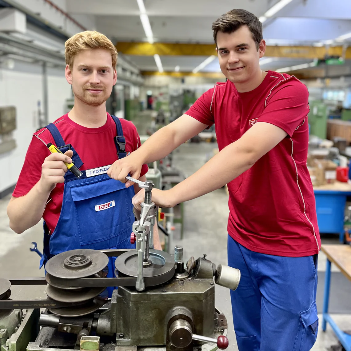 Two men in a workshop setting, both wearing red shirts and blue overalls. One is holding a screwdriver and the other is operating a machine tool with tesa tape nearby on the workbench. The background shows industrial equipment and a bright environment, highlighting the use of tesa tape in their projects. (This text has been generated by AI)