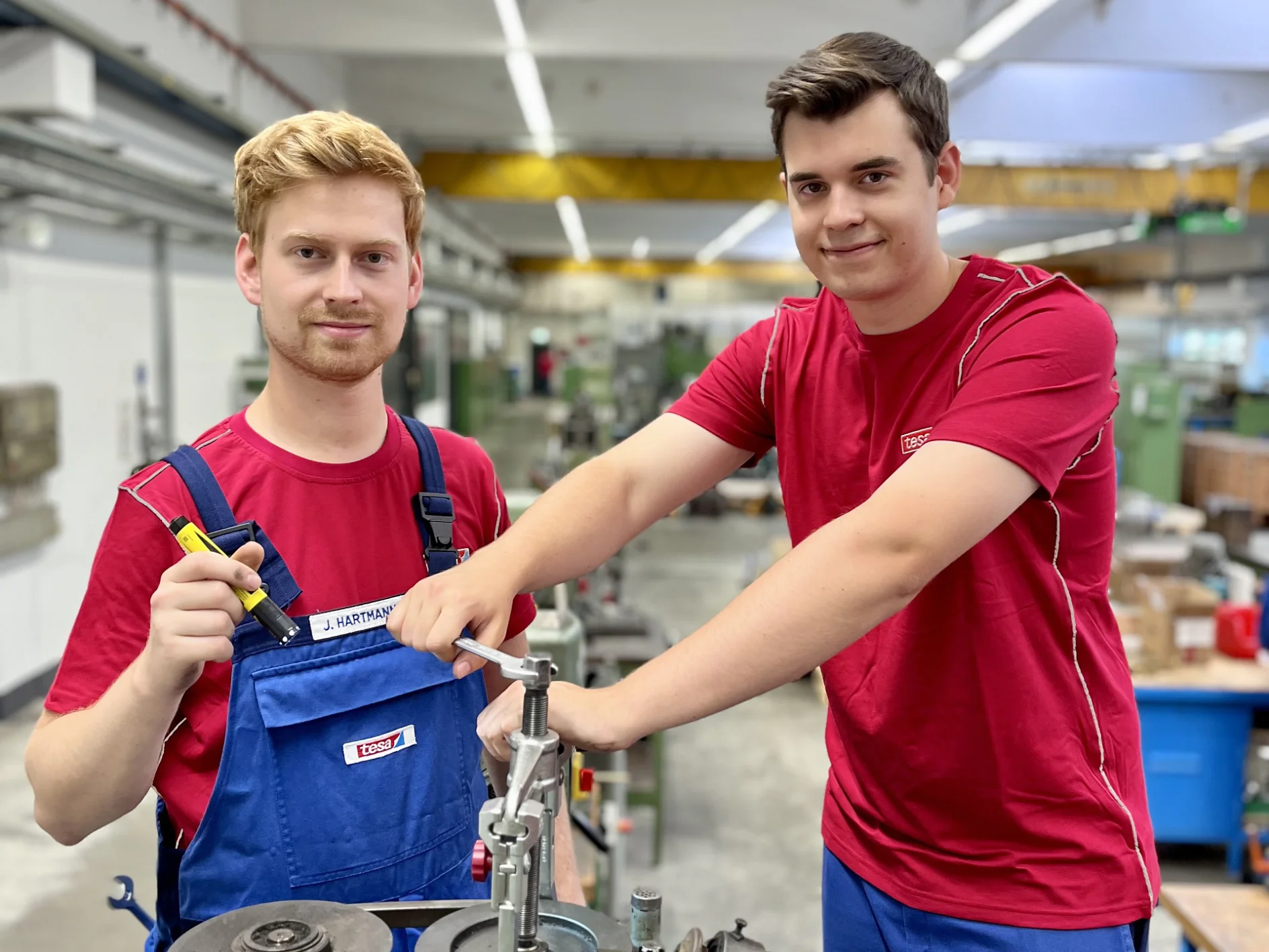 Two men in a workshop setting, both wearing red shirts and blue overalls. One is holding a screwdriver and the other is operating a machine tool with tesa tape nearby on the workbench. The background shows industrial equipment and a bright environment, highlighting the use of tesa tape in their projects. (This text has been generated by AI)