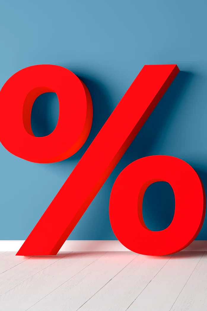 Ein großes rotes Prozentzeichen steht vor einer blauen Wand auf einem weißen Holzboden. Das Symbol ist mit Tesa-Klebeband befestigt und bleibt perfekt auf der linken Seite des Bildes positioniert. Dadurch bleibt auf der rechten Seite ein negativer Raum, wodurch ein minimalistischer und kräftiger visueller Effekt entsteht. (This text has been generated by AI)