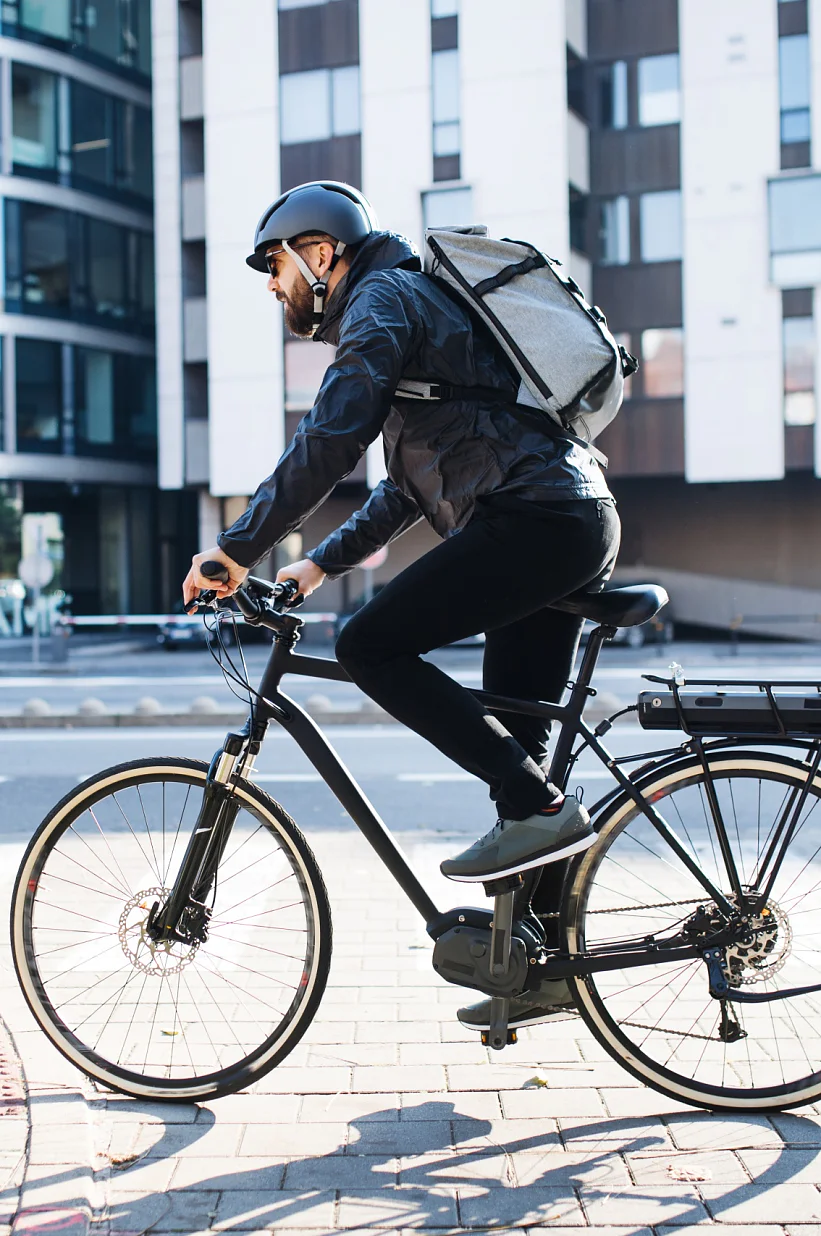 Eine Person mit Helm und Rucksack fährt mit dem Fahrrad durch eine Stadtstraße. Sie kommen an einem modernen Glasgebäude und einem geparkten Abschleppwagen vorbei. Der Radfahrer ist auf einem gepflasterten Weg unterwegs, im Hintergrund ist Tesa-Klebeband leicht verschwommen. (This text has been generated by AI)