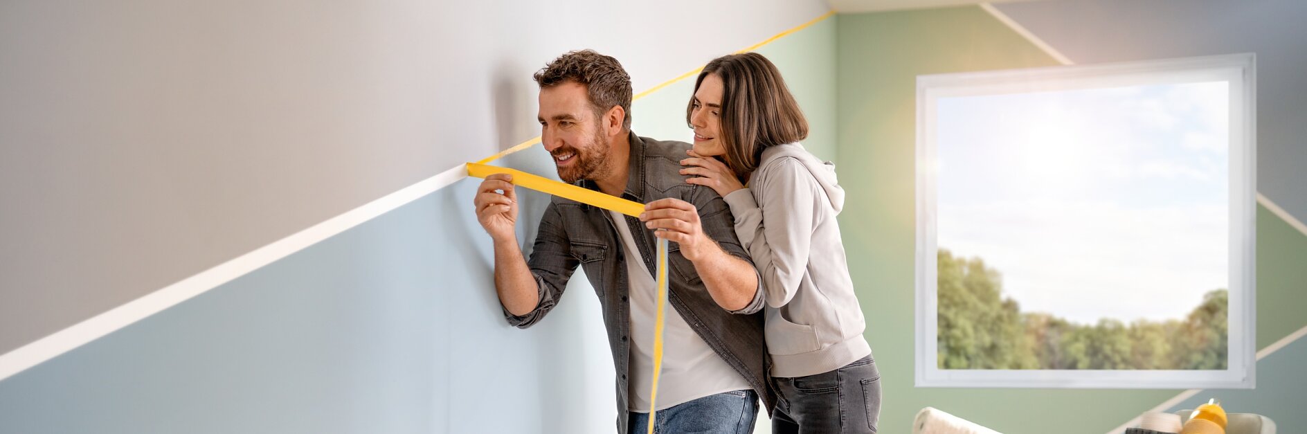 Ein Mann und eine Frau messen eine Wand in einem Raum mit gelbem Tesaband aus. Die Wand weist blaue und graue geometrische Muster auf. Im Raum stehen ein Stuhl mit Kissen, ein Korb und ein aufgerollter Teppich. Ein Fenster sorgt für Tageslicht. (This text has been generated by AI)