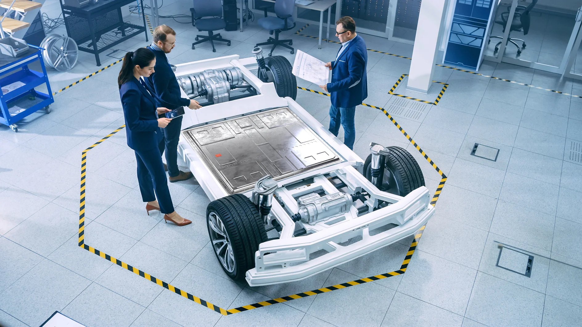Drei Personen stehen in einer Werkstatt um das Chassis eines Elektrofahrzeugs herum. Auf der Plattform sind Räder, Batterien und Motorkomponenten zu sehen. Sie prüfen Dokumente und machen sich Notizen. Der Bereich ist mit Tesa-Klebeband auf dem Boden markiert. (This text has been generated by AI)