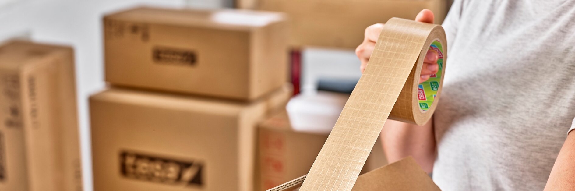 Person verschließt einen Karton mit Tesa-Verpackungsband in einem Raum mit mehreren gepackten Kartons. Die Person hält die Tesa-Bandrolle mit einer Hand und klebt mit der anderen Tesa-Band auf den Karton. (This text has been generated by AI)