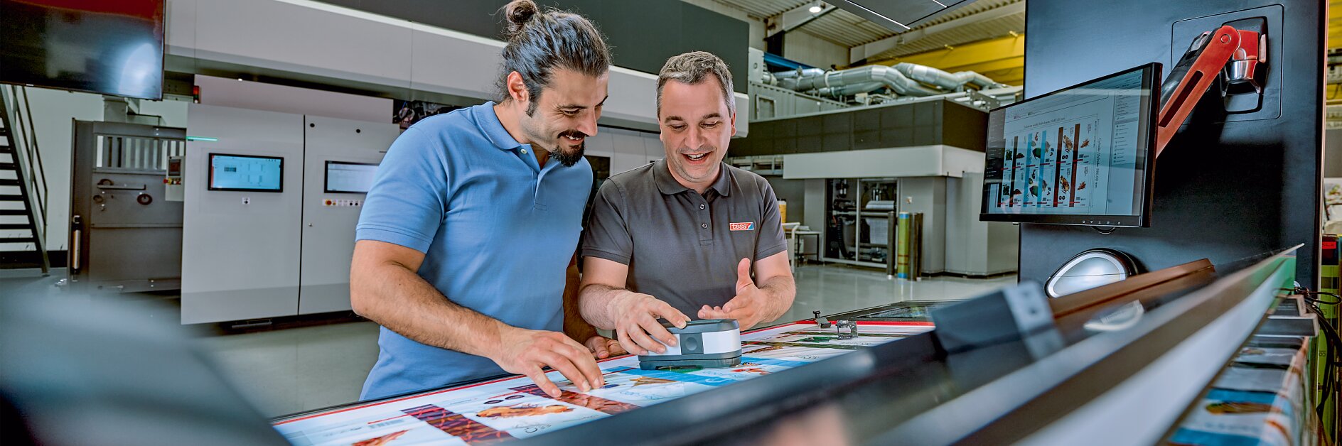Two men are standing in a modern printing facility, examining colorful printed materials on a large machine. One man is holding a small box of tesa tape and smiling. There are screens and equipment in the background.