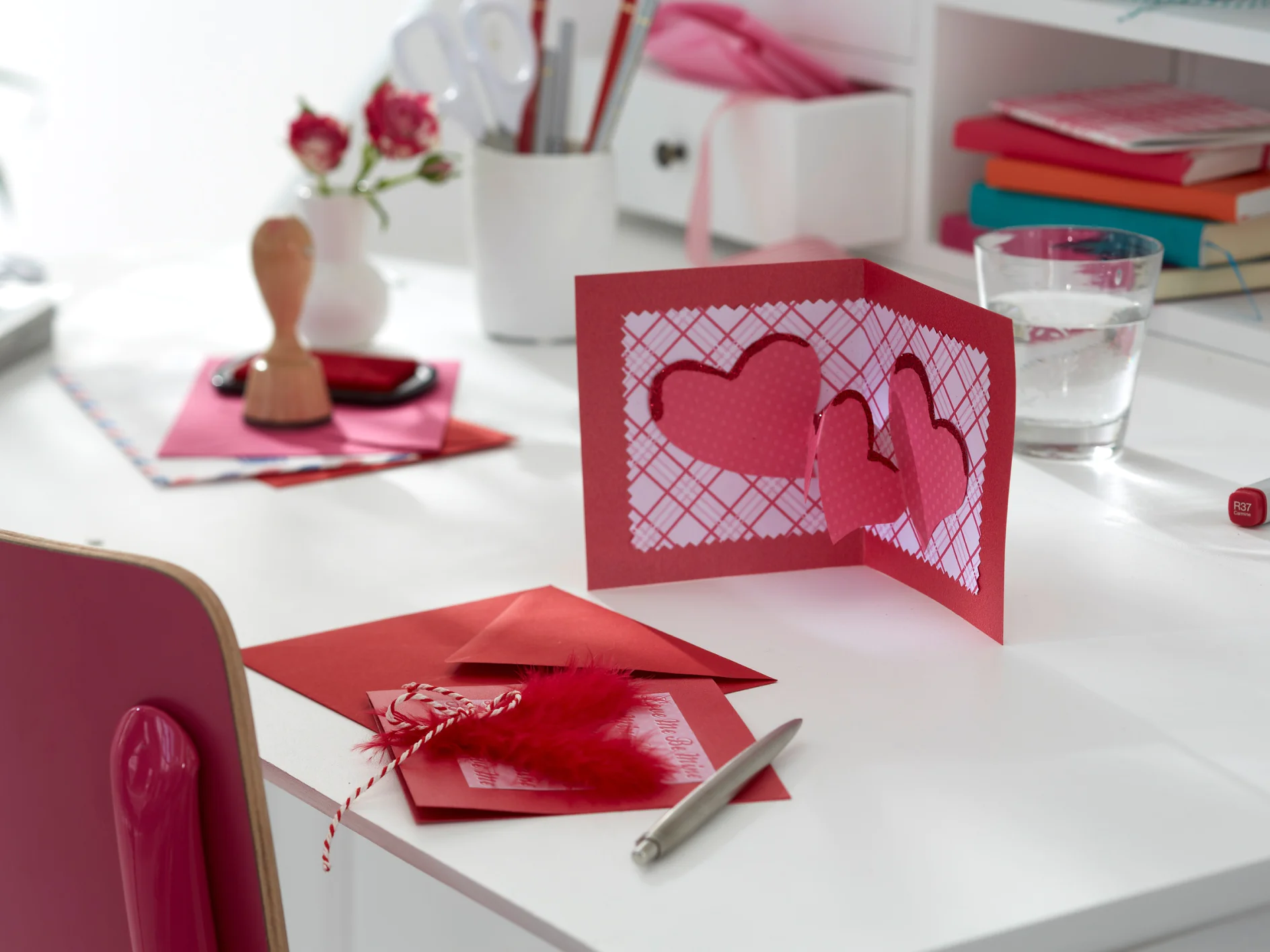 Eine rote Valentinstagskarte mit ausgeschnittenen Herzen liegt auf einem weißen Schreibtisch. Daneben liegen ein roter Umschlag, ein Stift, eine rote Feder, Buntstifte, Bücher, ein Glas Wasser und kleine Vasen mit roten Blumen. Auf dem Schreibtisch liegt eine Rolle Tesa-Klebeband, um die einzelnen Teile zusammenzuhalten. Ein roter Stuhl ist teilweise sichtbar. (This text has been generated by AI)