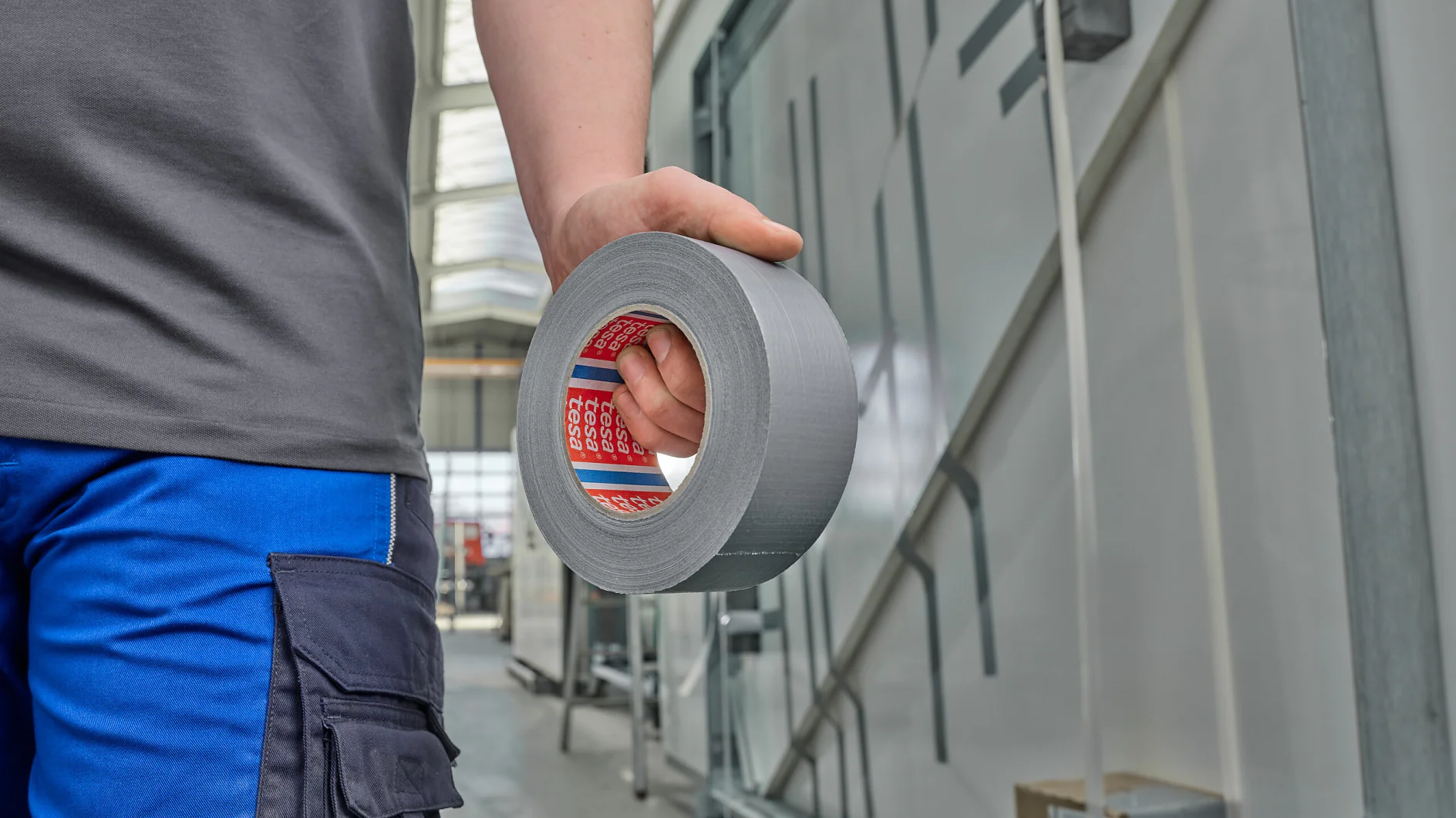 person in blue pants holding a roll of silver tesa duct tape in an industrial indoor setting (This text has been generated by AI)