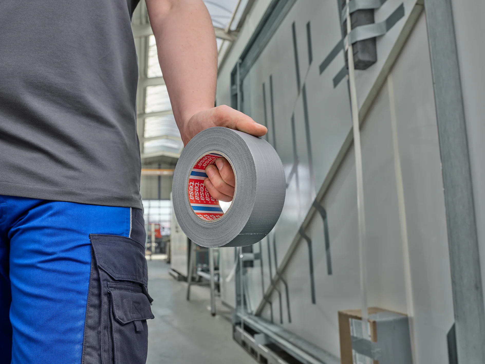 person in blue pants holding a roll of silver tesa duct tape in an industrial indoor setting (This text has been generated by AI)