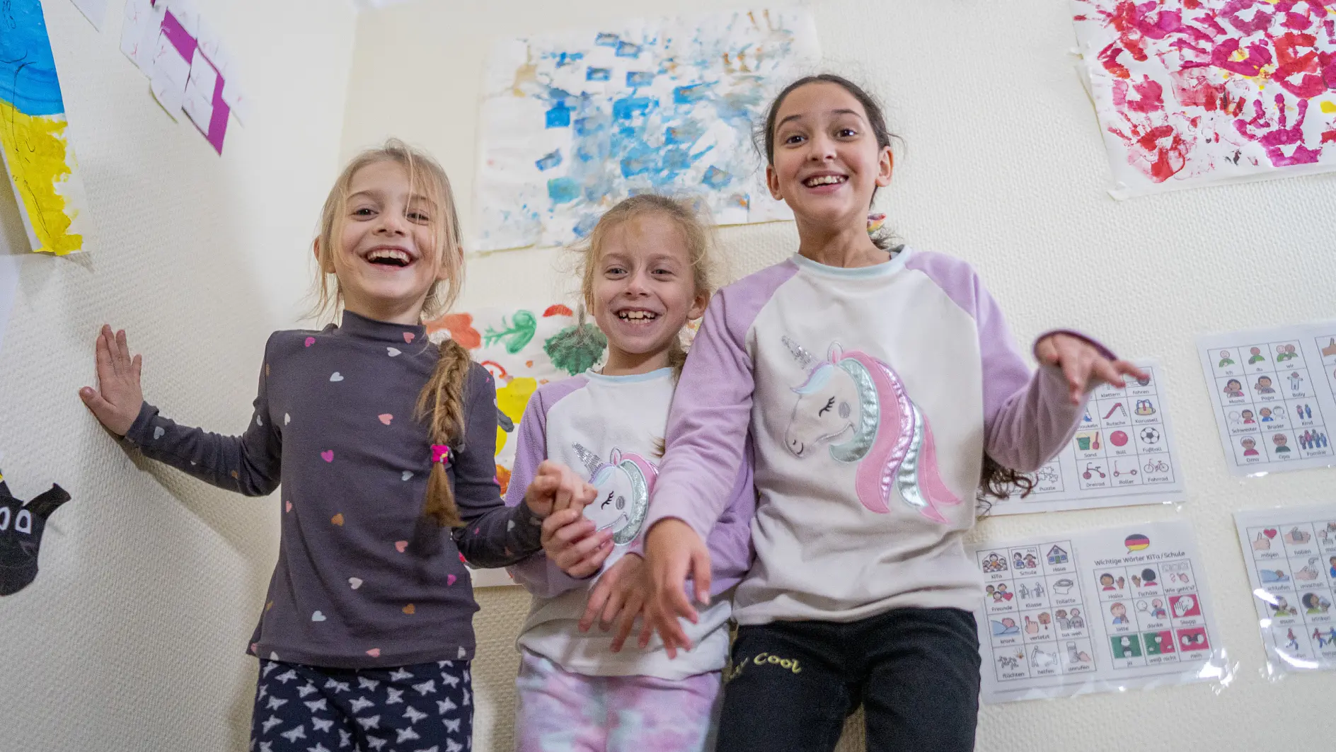 Three young girls stand together smiling in a room with colorful artwork on the walls. They are wearing casual clothes, and their expressions are joyful. The decor features various colorful paintings and educational posters, all meticulously secured to the walls using tesa tape. (This text has been generated by AI)