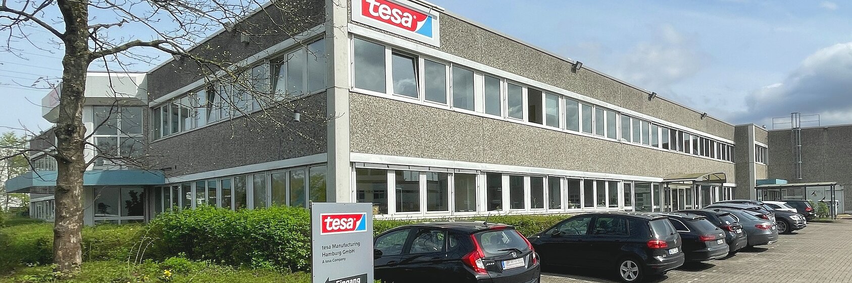Das Bild zeigt ein zweistöckiges Bürogebäude mit einem Schild mit dem tesa-Logo. Davor parken mehrere Autos, links ist ein Baum zu sehen. Der Himmel ist teilweise bewölkt. (This text has been generated by AI)