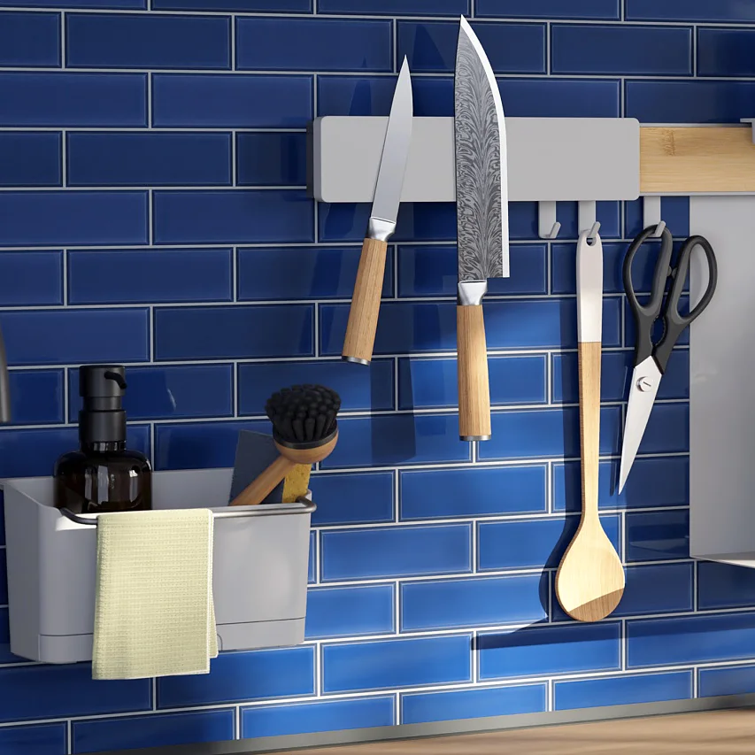 Eine Küchenrückwand mit blauen U-Bahn-Fliesen zeigt verschiedene Utensilien und Gegenstände. Messer, ein Löffel und eine Bürste hängen an einem Gestell. Eine Topfpflanze, eine Flasche und ein Papierhandtuchhalter sind ebenfalls vorhanden. Im Vordergrund steht ein schwarzer Wasserhahn. Alle Gegenstände sind für dauerhaften Halt und Zuverlässigkeit sicher mit Tesa-Klebeband befestigt. (This text has been generated by AI)