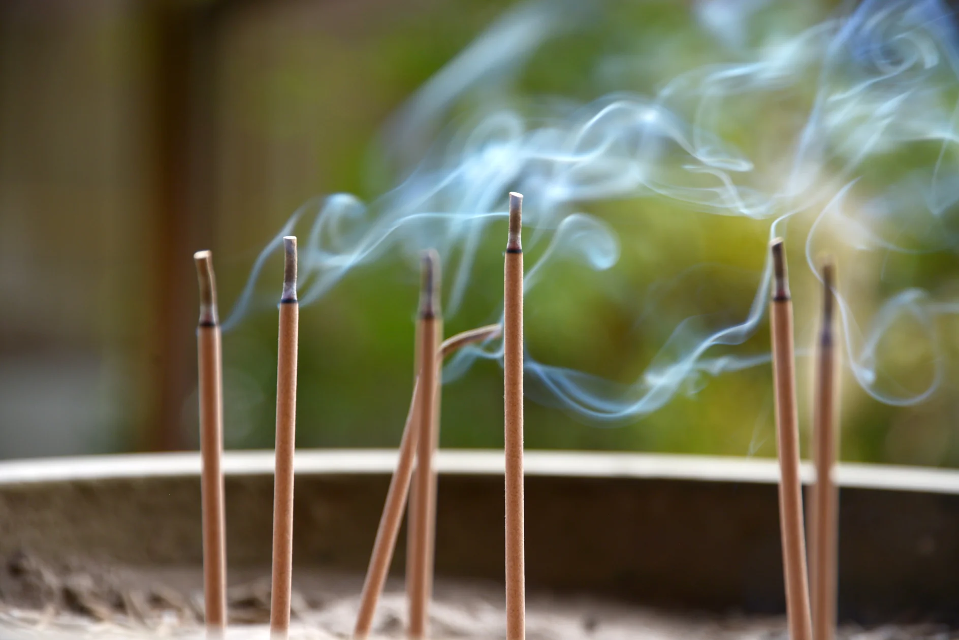 Mehrere Räucherstäbchen brennen im Sand und geben dünne Rauchschwaden ab. Der Hintergrund ist sanft verschwommen, wodurch Rauch und Weihrauch hervorgehoben werden. Die Szene vermittelt eine friedliche und meditative Atmosphäre. (This text has been generated by AI)