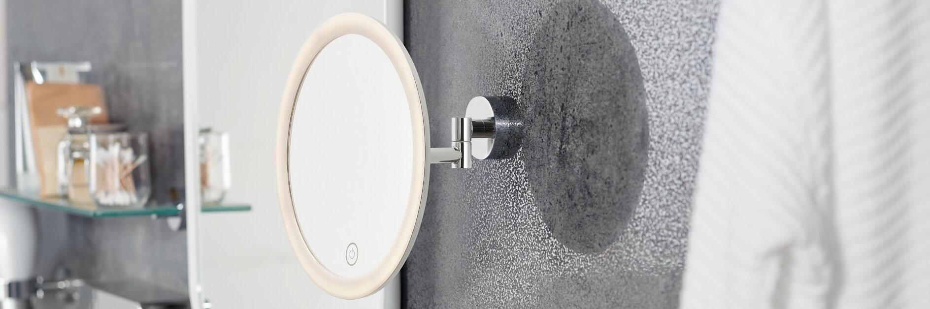 Ein modernes Badezimmer verfügt über einen runden, beleuchteten Spiegel, der mit Tesa-Klebeband an einer strukturierten grauen Wand befestigt ist. Rechts hängt ein Bademantel und auf Regalen stehen verschiedene Toilettenartikel und Glasbehälter. Im Vordergrund ist das Waschbecken zu sehen. (This text has been generated by AI)
