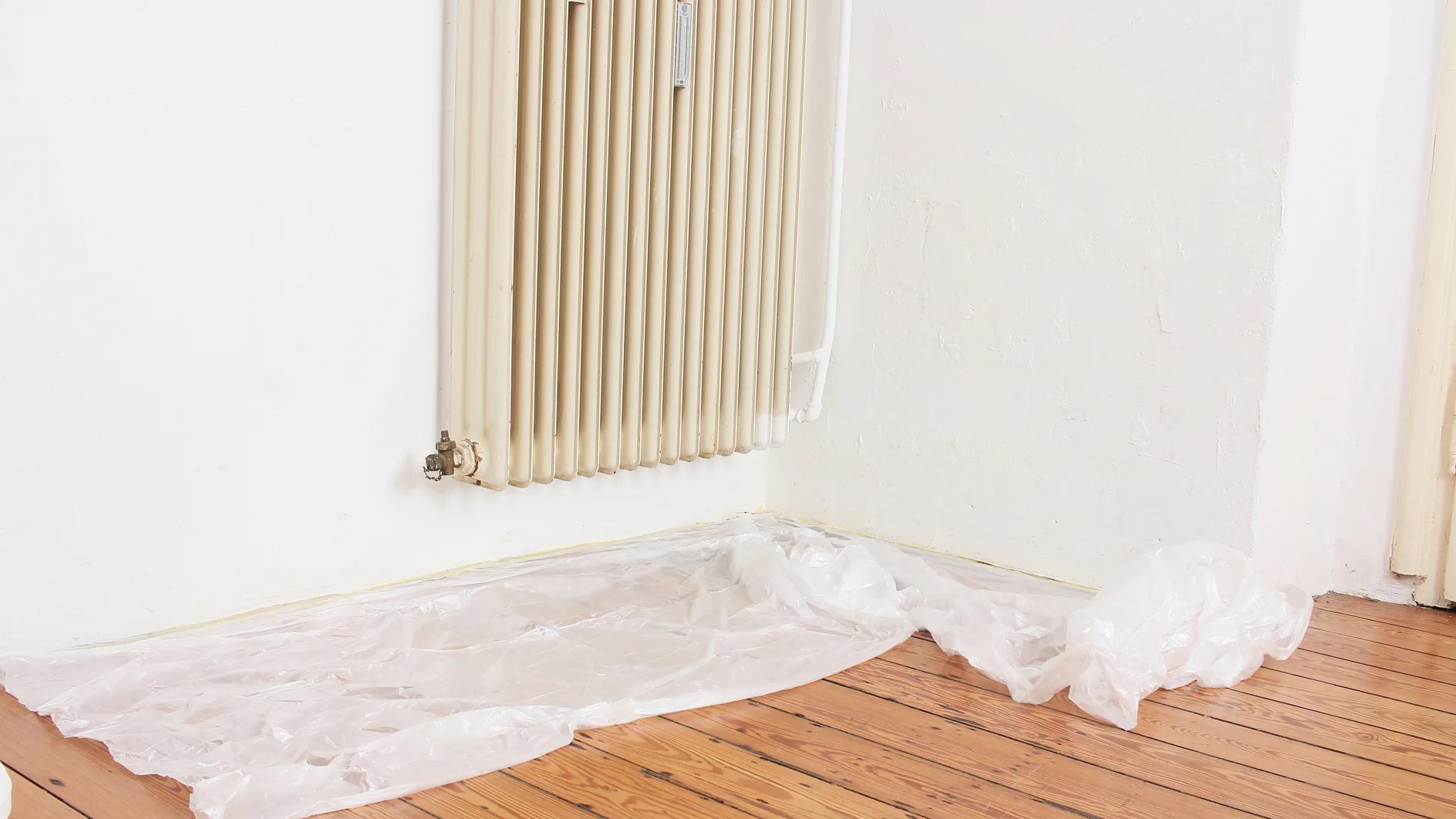 An einer weißen Wand ist ein cremefarbener Heizkörper montiert. Auf dem darunterliegenden Holzboden liegt ein Stück transparentes Tesa-Klebeband, das vom Heizkörperfuß die Wand entlang verläuft. (This text has been generated by AI)