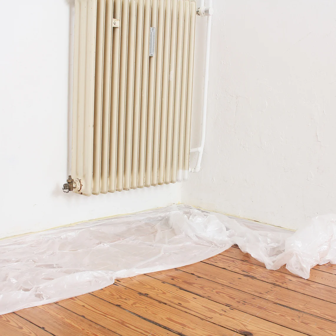 An einer weißen Wand ist ein cremefarbener Heizkörper montiert. Auf dem darunterliegenden Holzboden liegt ein Stück transparentes Tesa-Klebeband, das vom Heizkörperfuß die Wand entlang verläuft. (This text has been generated by AI)
