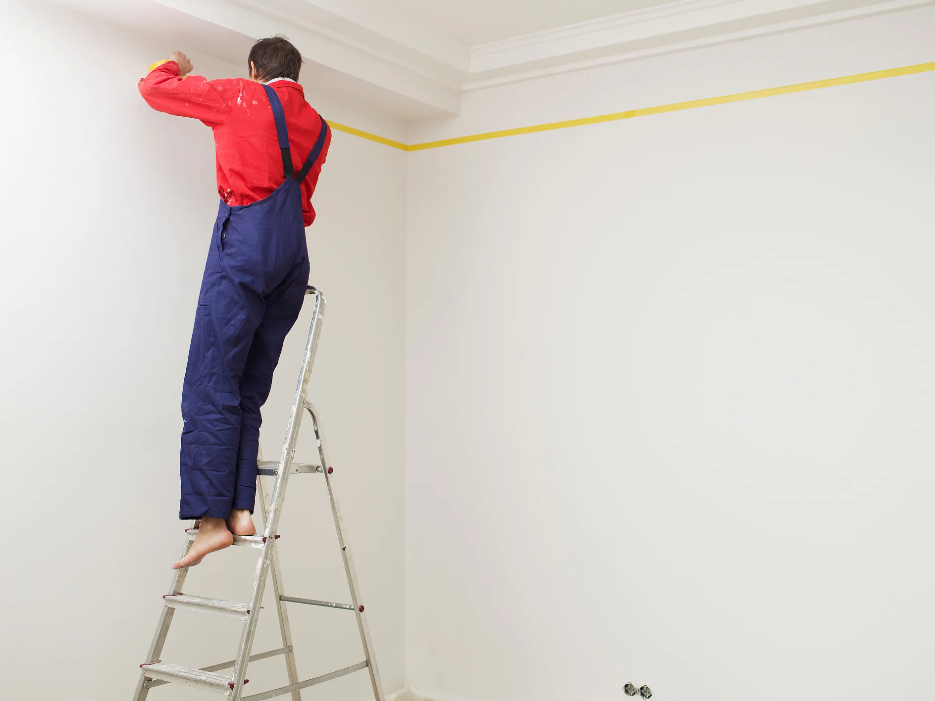 Eine Person in rotem Hemd und blauem Overall steht barfuß auf einer Metallleiter und streicht eine weiße Wand. Eine gelbe Linie Tesa-Klebeband markiert die Oberkante der Wand nahe der Decke. In Bodennähe sind zwei kleine Vorrichtungen zu sehen. (This text has been generated by AI)