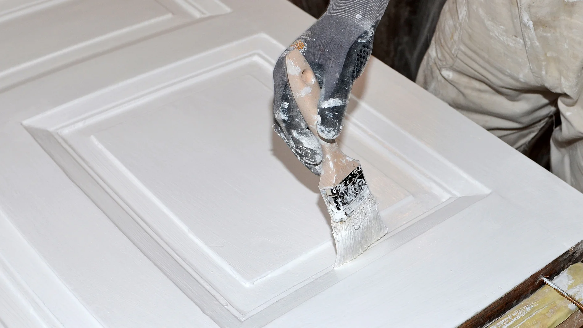 Eine Person mit Handschuhen streicht eine Holztür mit einem Pinsel weiß. Die Tür hat erhabene rechteckige Paneele, und Farbe ist auf dem Handschuh und dem Pinsel sichtbar. Die Person ist dabei, die Oberfläche gleichmäßig mit Tesaband abzudecken. (This text has been generated by AI)