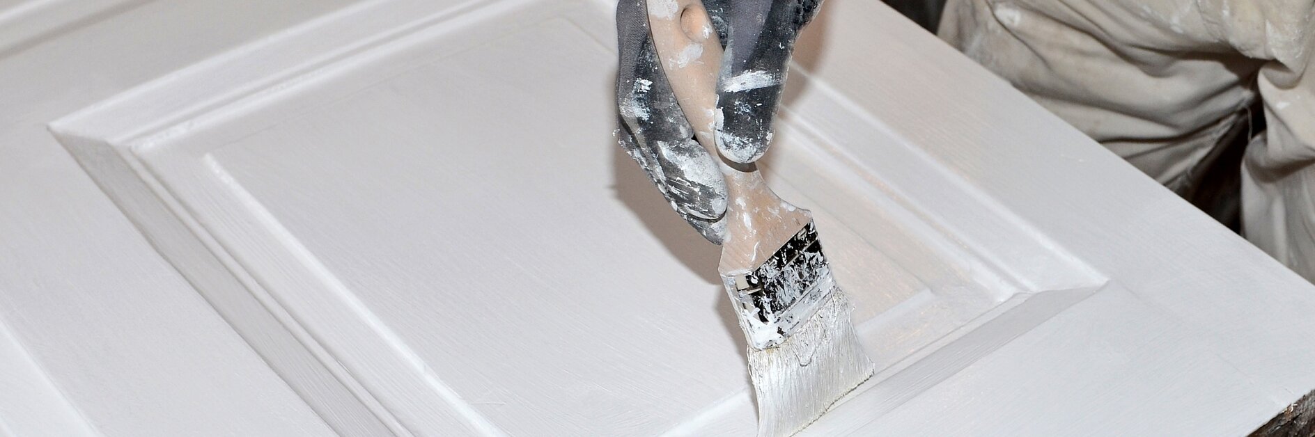 Eine Person mit Handschuhen streicht eine Holztür mit einem Pinsel weiß. Die Tür hat erhabene rechteckige Paneele, und Farbe ist auf dem Handschuh und dem Pinsel sichtbar. Die Person ist dabei, die Oberfläche gleichmäßig mit Tesaband abzudecken. (This text has been generated by AI)