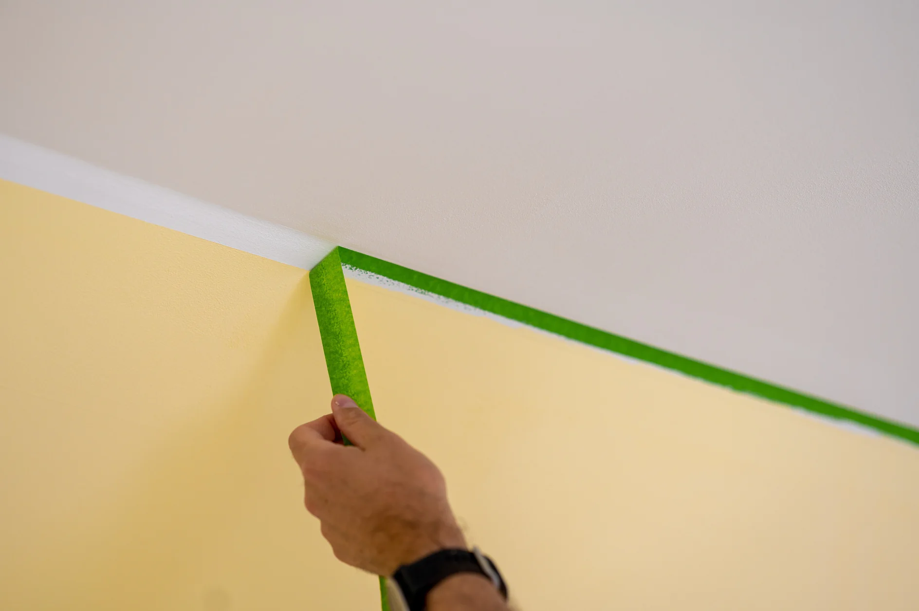 Painter removes masking tape and creates a sharp border between a yellow and white painted part of a wall.