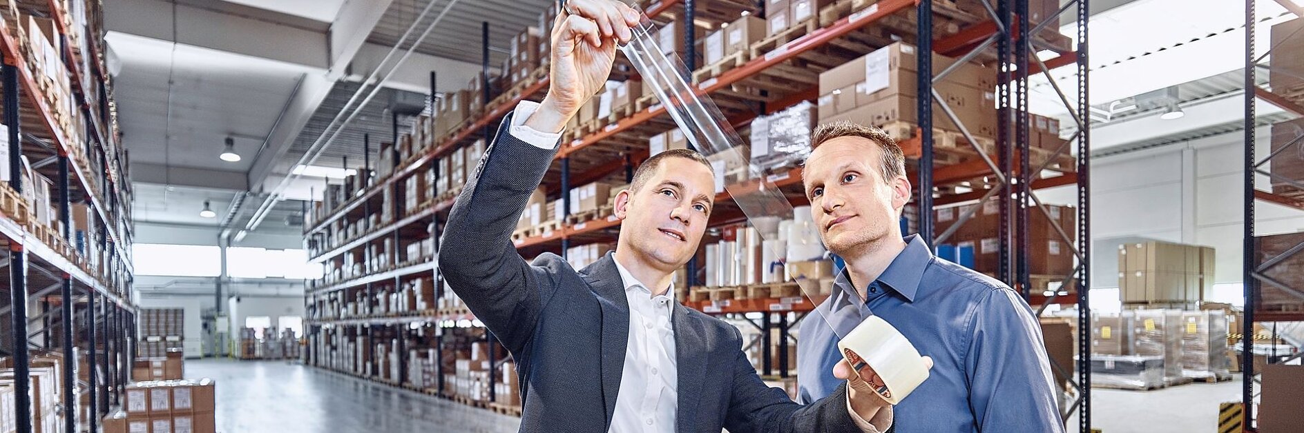 Zwei Männer in Businesskleidung stehen in einer Lagerhalle. Einer hält eine Rolle Tesa-Klebeband in der Hand, der andere betrachtet einen Streifen davon und hält ihn gegen das Licht. Im Hintergrund sind Regale voller Kartons und Pakete zu sehen. (This text has been generated by AI)