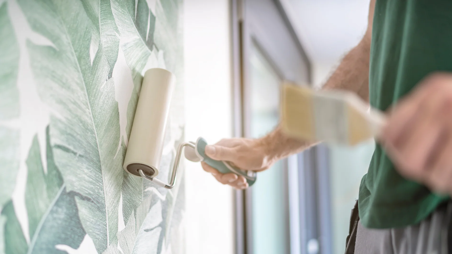 Eine Person bringt mit einer Rolle eine Tapete an einer Wand mit einem grünen Blättermuster an. Der Fokus liegt auf dem Tapezieren, im Vordergrund ist ein unscharfes Tesa-Band zu sehen. Natürliches Licht fällt durch ein Fenster auf der rechten Seite ein. (This text has been generated by AI)