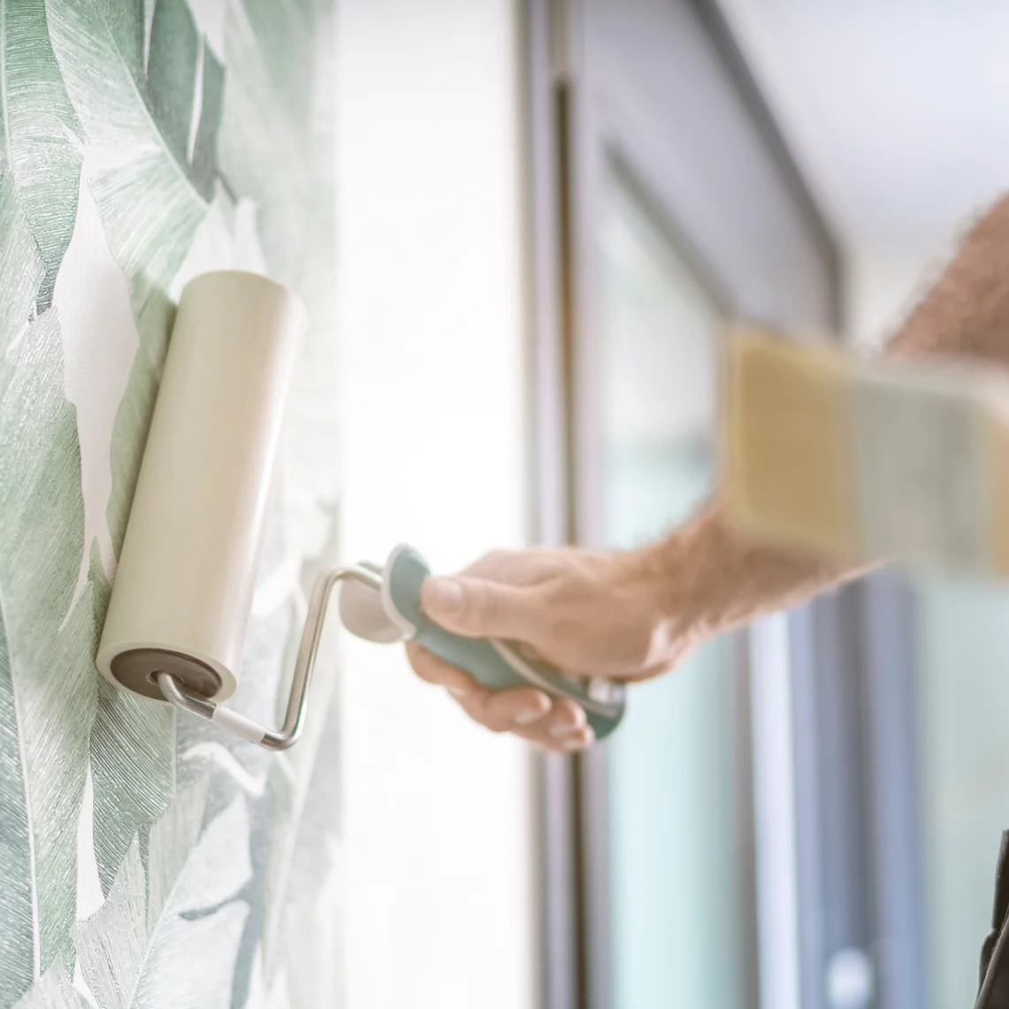 Eine Person bringt mit einer Rolle eine Tapete an einer Wand mit einem grünen Blättermuster an. Der Fokus liegt auf dem Tapezieren, im Vordergrund ist ein unscharfes Tesa-Band zu sehen. Natürliches Licht fällt durch ein Fenster auf der rechten Seite ein. (This text has been generated by AI)