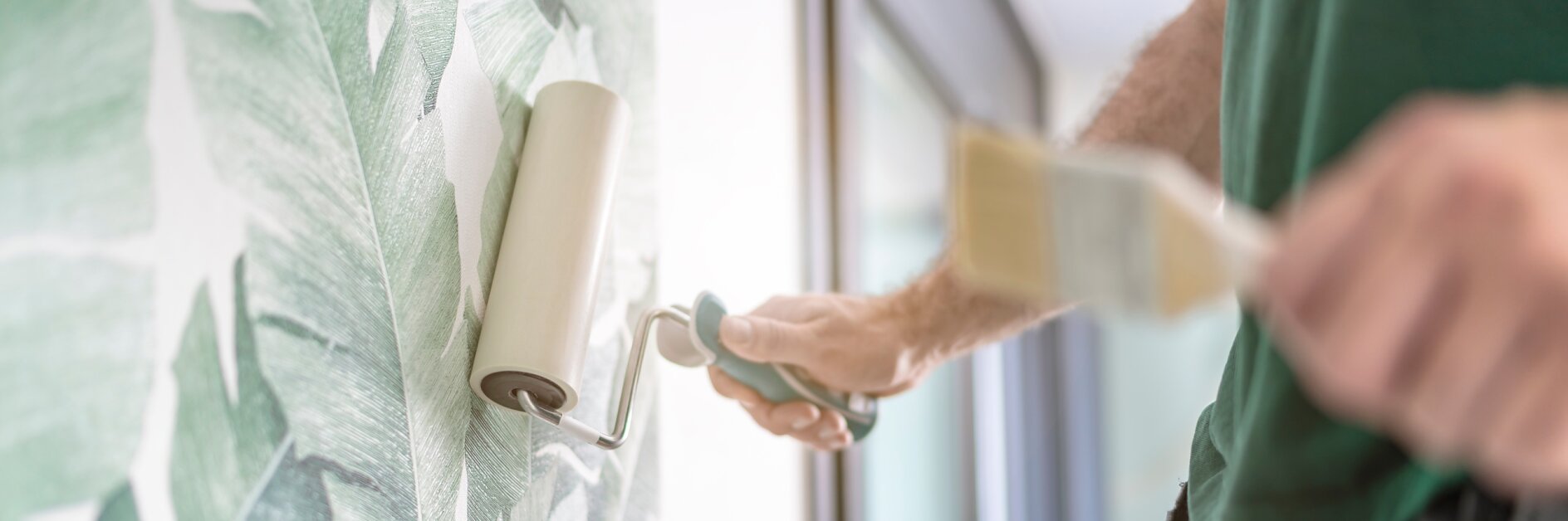 Eine Person bringt mit einer Rolle eine Tapete an einer Wand mit einem grünen Blättermuster an. Der Fokus liegt auf dem Tapezieren, im Vordergrund ist ein unscharfes Tesa-Band zu sehen. Natürliches Licht fällt durch ein Fenster auf der rechten Seite ein. (This text has been generated by AI)