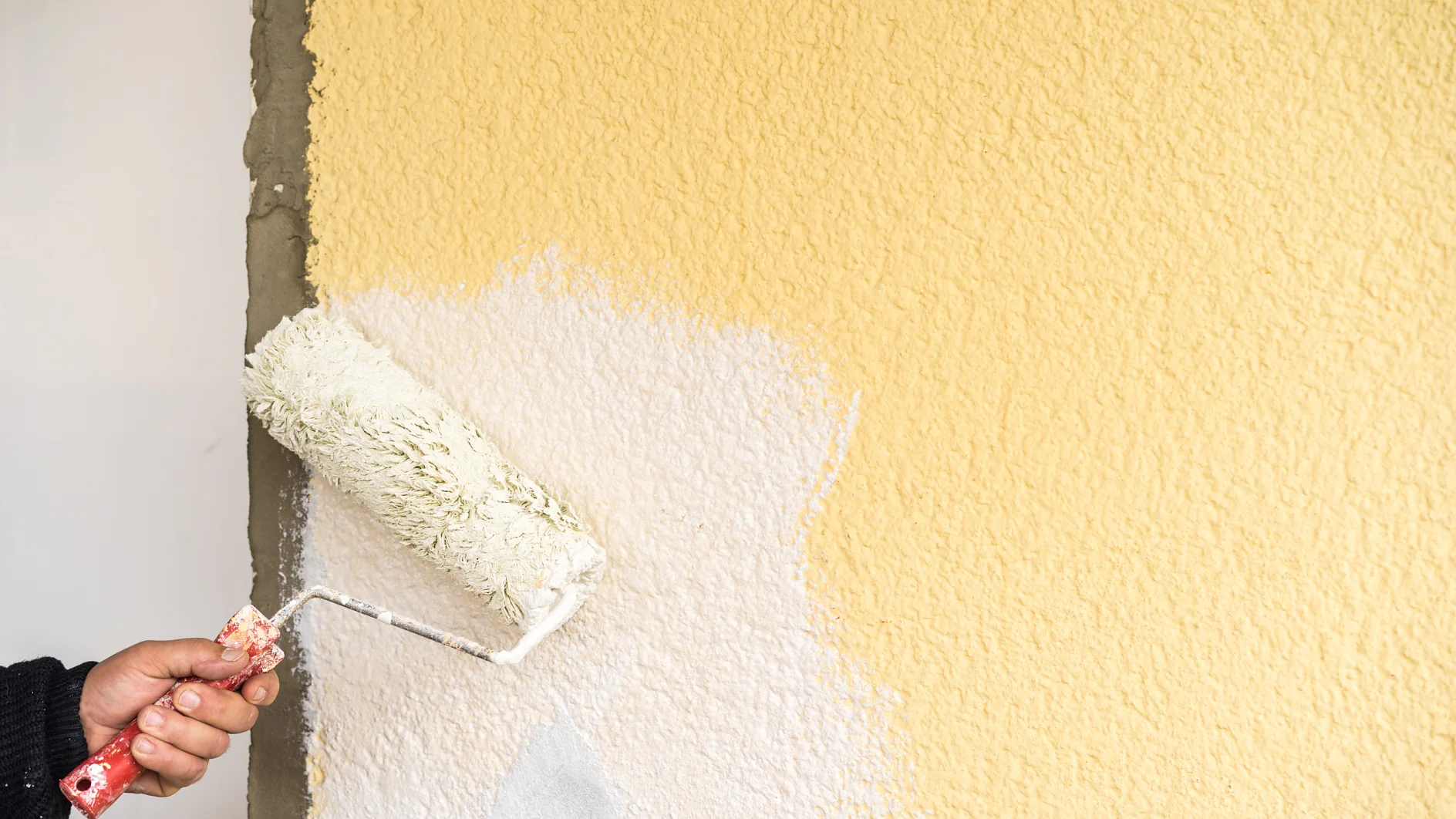 Eine Person streicht eine strukturierte Wand mit einer Farbrolle. Die Wand ist teilweise mit weißer Farbe bedeckt, die zuvor einen gelben Anstrich hatte. Die Hand, die die Rolle hält, ist sichtbar, und das Tesa-Klebeband, mit dem bestimmte Bereiche abgedeckt wurden, hilft dabei, saubere Kanten und scharfe Linien auf der gestrichenen Oberfläche zu erzeugen, während die Farbwanne außer Sicht bleibt. (This text has been generated by AI)