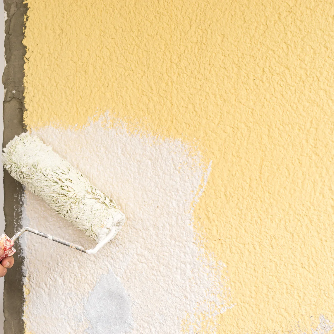 Eine Person streicht eine strukturierte Wand mit einer Farbrolle. Die Wand ist teilweise mit weißer Farbe bedeckt, die zuvor einen gelben Anstrich hatte. Die Hand, die die Rolle hält, ist sichtbar, und das Tesa-Klebeband, mit dem bestimmte Bereiche abgedeckt wurden, hilft dabei, saubere Kanten und scharfe Linien auf der gestrichenen Oberfläche zu erzeugen, während die Farbwanne außer Sicht bleibt. (This text has been generated by AI)