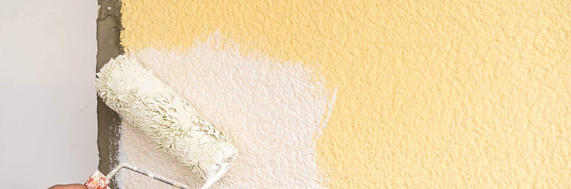 Eine Person streicht eine strukturierte Wand mit einer Farbrolle. Die Wand ist teilweise mit weißer Farbe bedeckt, die zuvor einen gelben Anstrich hatte. Die Hand, die die Rolle hält, ist sichtbar, und das Tesa-Klebeband, mit dem bestimmte Bereiche abgedeckt wurden, hilft dabei, saubere Kanten und scharfe Linien auf der gestrichenen Oberfläche zu erzeugen, während die Farbwanne außer Sicht bleibt. (This text has been generated by AI)