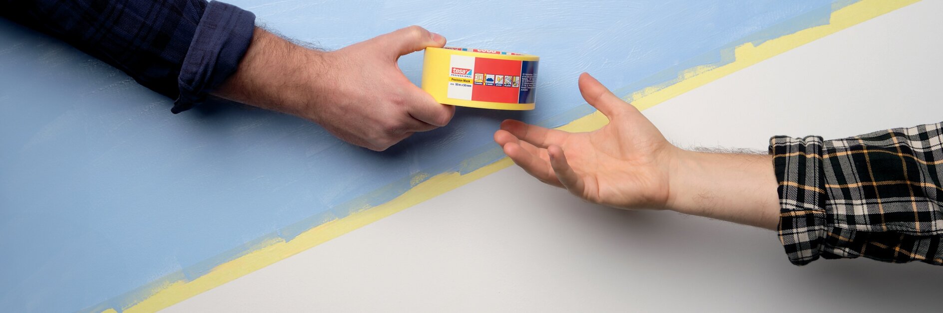 Vor einem Hintergrund mit einer diagonalen blau-weißen Wandteilung sind zwei Hände zu sehen. Die Hand, die von links kommt, hält eine kleine Schachtel mit Buntstiften, während die andere Hand, die von rechts kommt, im Begriff ist, sie zu ergreifen. Beide Personen sind in karierte Hemden gekleidet. (This text has been generated by AI)