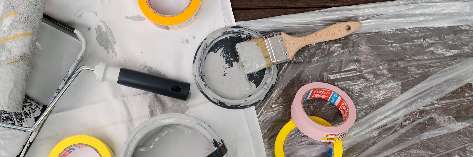 Ein Flachbild von Malutensilien auf einer Holzoberfläche. Enthält eine Farbrolle mit einer Schale, eine offene graue Farbdose, einen Pinsel und mehrere Rollen Tesa-Klebeband in Gelb, Rosa und Orange. Darunter befinden sich ein weißes Tuch und eine Plastikfolie. (This text has been generated by AI)