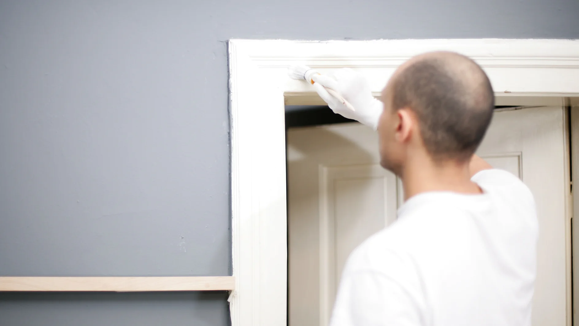 Eine Person mit einem weißen Handschuh streicht mit Tesa-Klebeband den Türrahmen vor einer grauen Wand weiß. Sie benutzt einen kleinen Pinsel und trägt ein weißes T-Shirt. Die Tür ist offen und gibt den Blick auf eine weitere weiße Tür im Hintergrund frei. (This text has been generated by AI)
