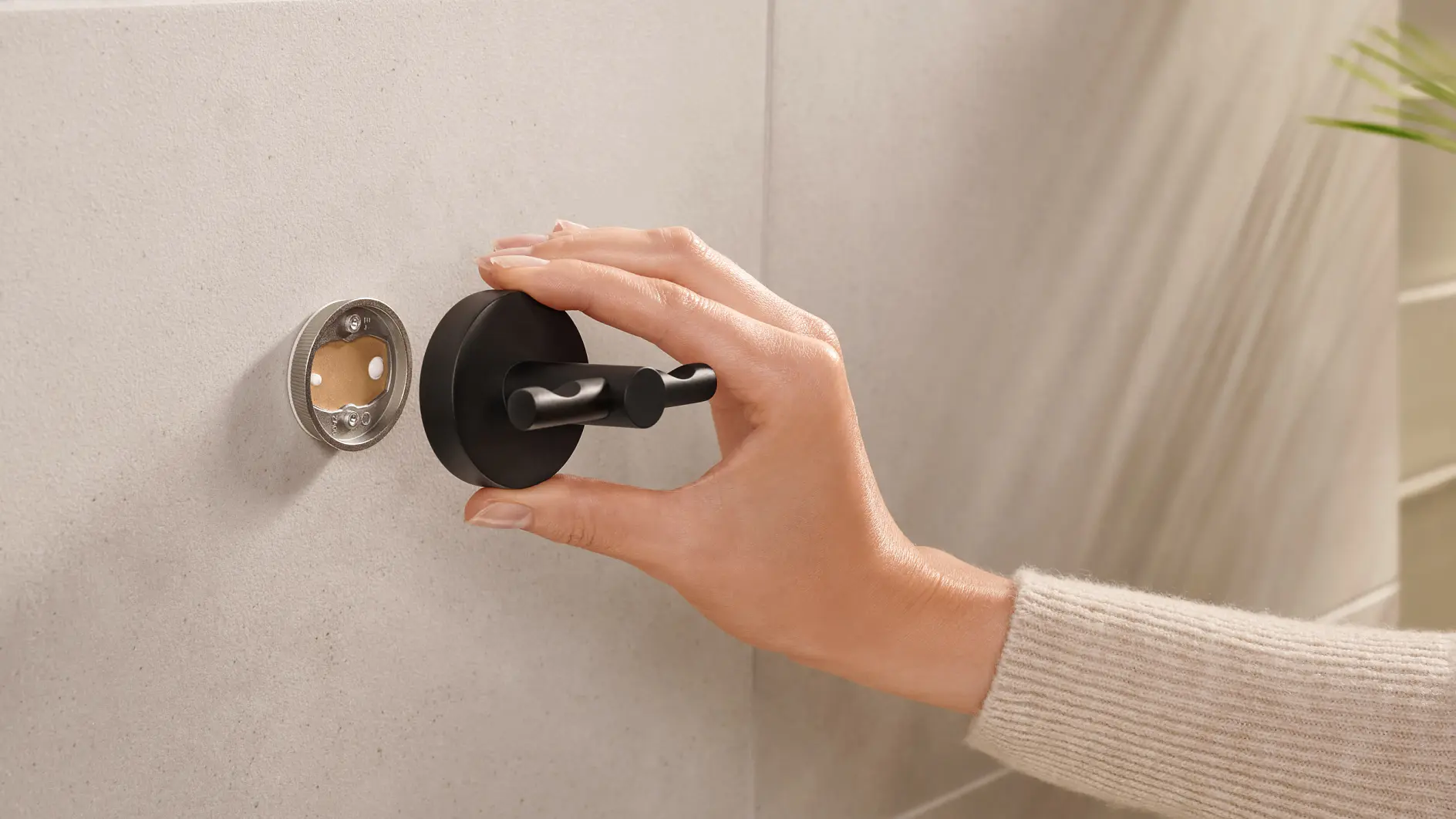 A hand wearing a beige sweater is adjusting a black wall-mounted hook on a light-colored tiled wall using tesa tape. The hand is holding the hook as it aligns with an open circular fitting on the wall. (This text has been generated by AI)