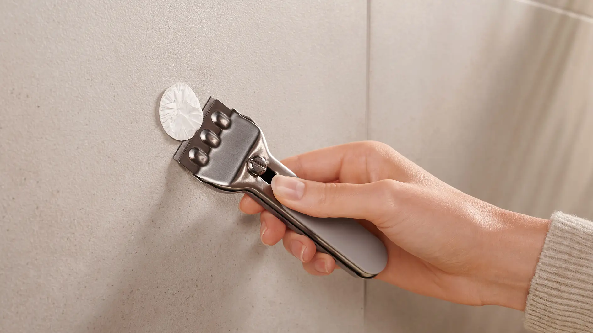 A persons hand holding a metal tool with a grey handle, pressing it against a circular metallic object on a smooth, light-colored vertical surface using tesa tape. (This text has been generated by AI)