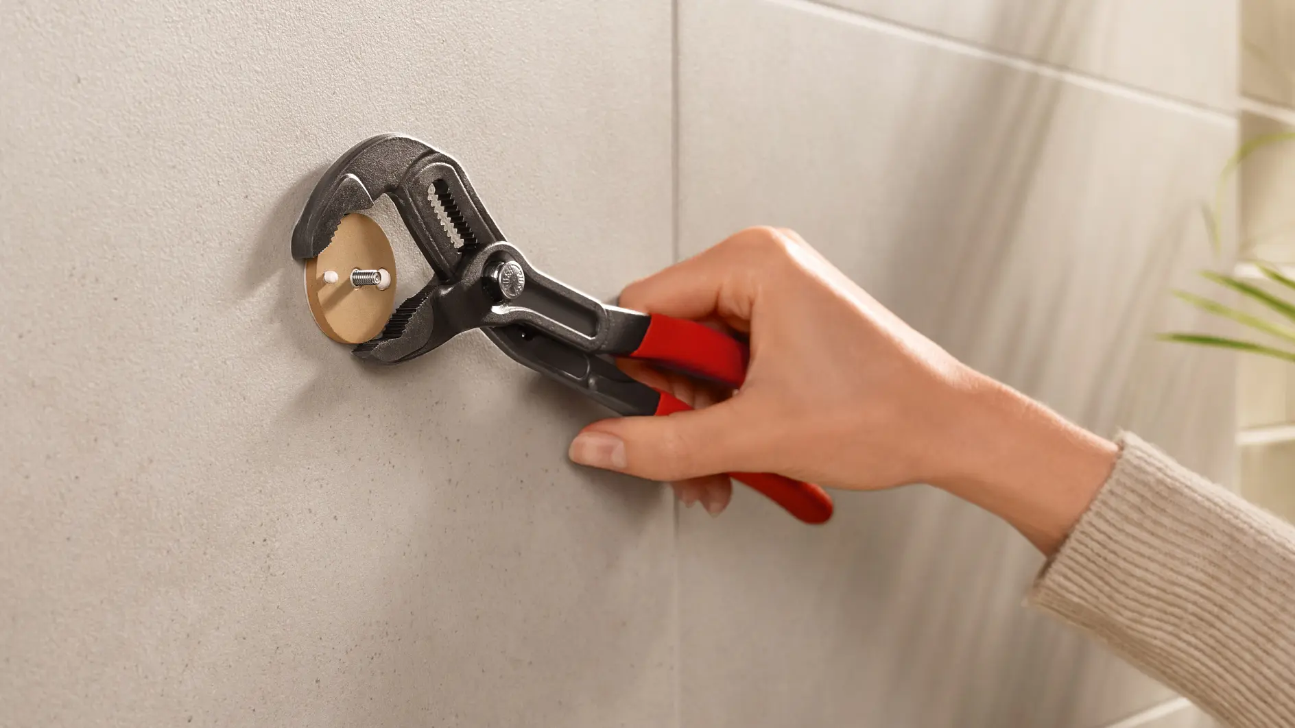 A person is using an adjustable wrench with red handles to tighten or loosen a round fitting on a wall. The wall appears to be tiled in a light gray color, and there are some blurred green leaves visible on the side. (This text has been generated by AI)