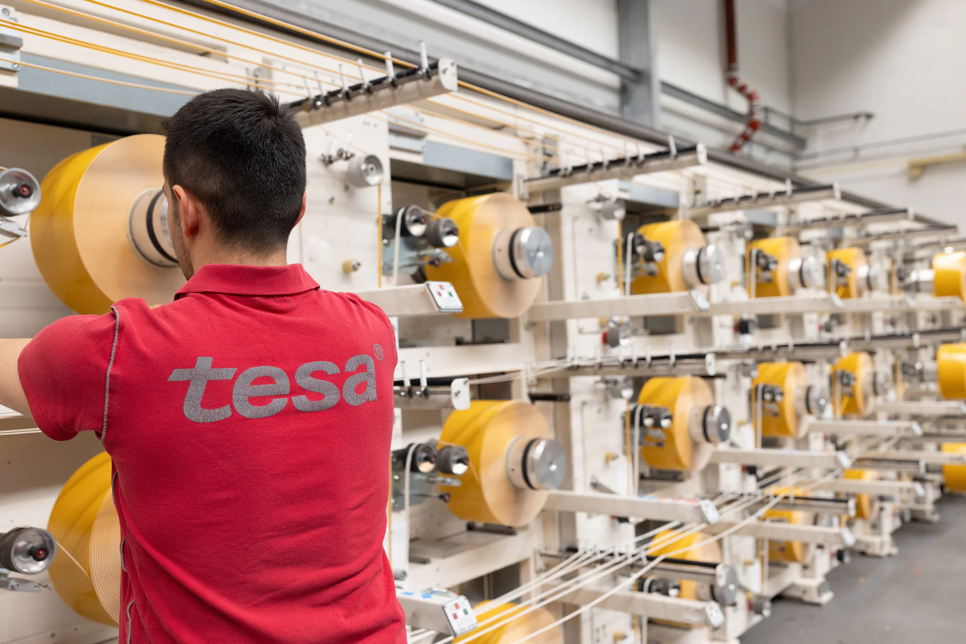 A person in a red shirt with the logo tesa works in a factory. They are facing several large spools of yellow tesa tape on a production line. The environment appears industrial, with metal structures and machinery. (This text has been generated by AI)