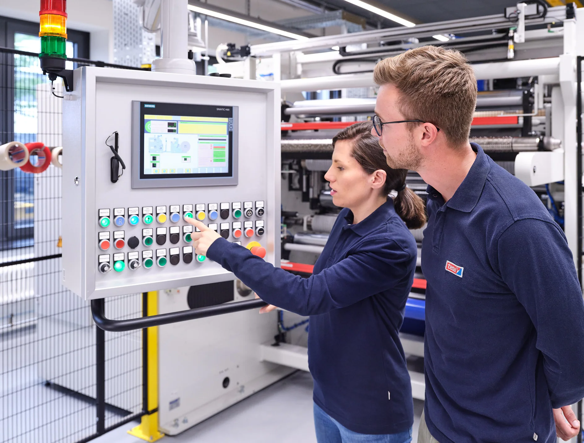 Two people in blue shirts are standing in front of a control panel with multiple buttons and a digital display. One person is pointing at the screen, while the other observes. tesa tape is used throughout the machinery visible in the background. (This text has been generated by AI)