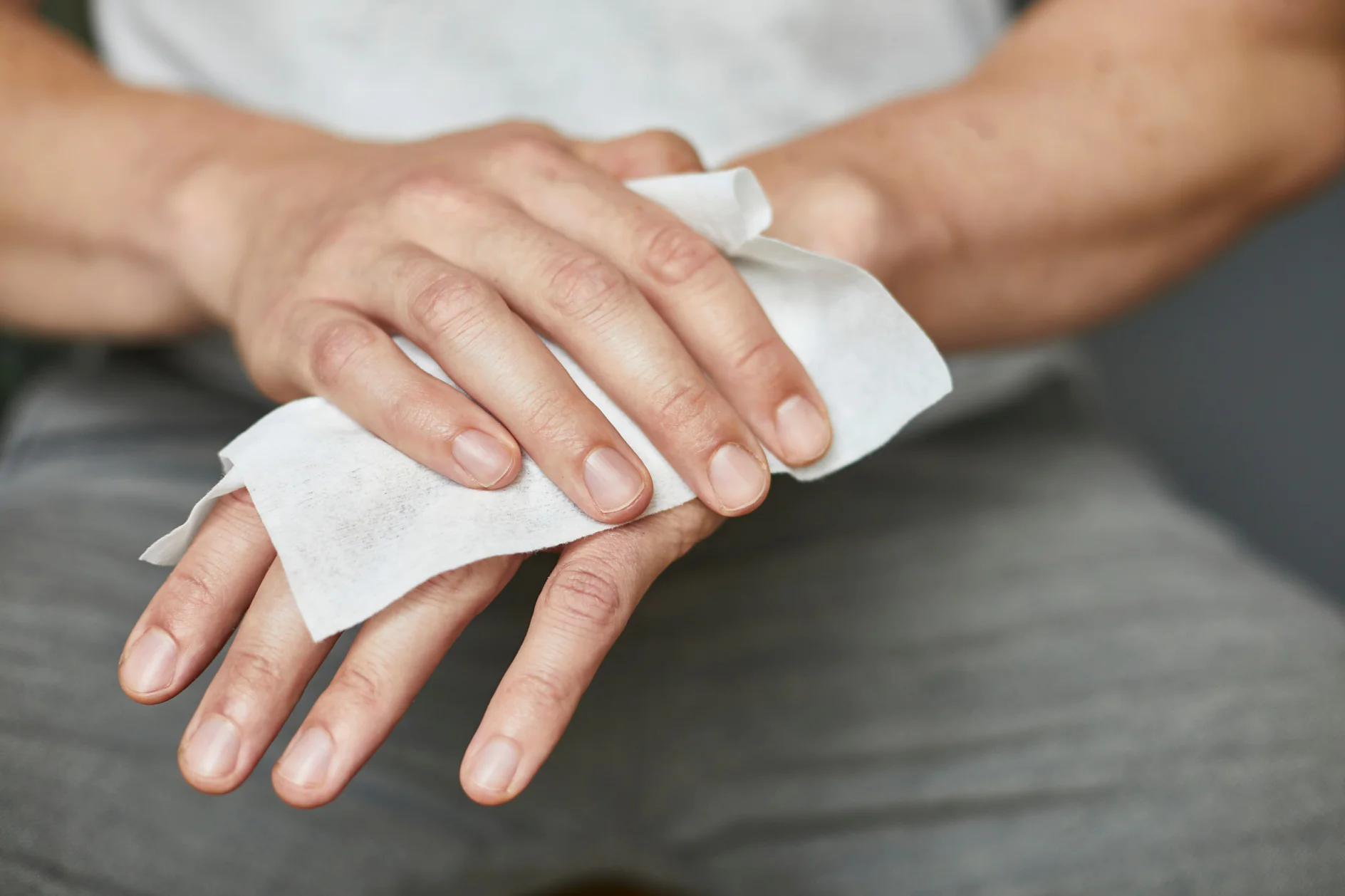 Eine Person reinigt ihre linke Hand mit einem weißen Feuchttuch. Die rechte Hand der Person hält das Tesaband, während die linke Hand abgewischt wird. Die Person trägt eine graue Hose und ein helles Hemd. Der Fokus liegt auf den Händen und dem Tuch. (This text has been generated by AI)
