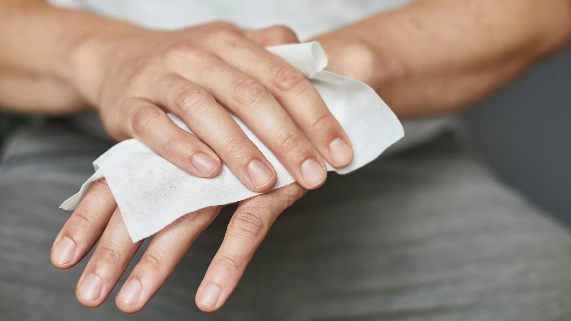 Eine Person reinigt ihre linke Hand mit einem weißen Feuchttuch. Die rechte Hand der Person hält das Tesaband, während die linke Hand abgewischt wird. Die Person trägt eine graue Hose und ein helles Hemd. Der Fokus liegt auf den Händen und dem Tuch. (This text has been generated by AI)