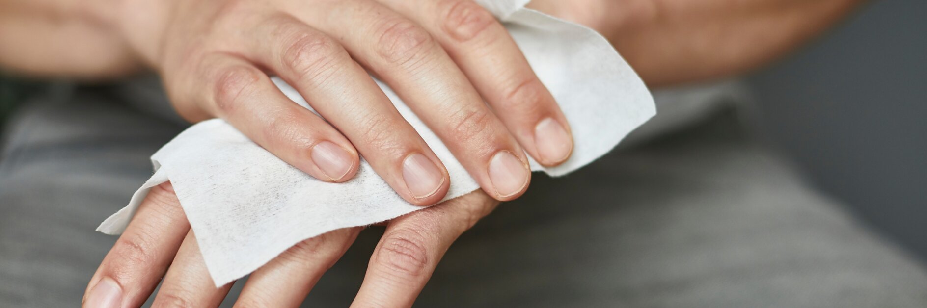 Eine Person reinigt ihre linke Hand mit einem weißen Feuchttuch. Die rechte Hand der Person hält das Tesaband, während die linke Hand abgewischt wird. Die Person trägt eine graue Hose und ein helles Hemd. Der Fokus liegt auf den Händen und dem Tuch. (This text has been generated by AI)