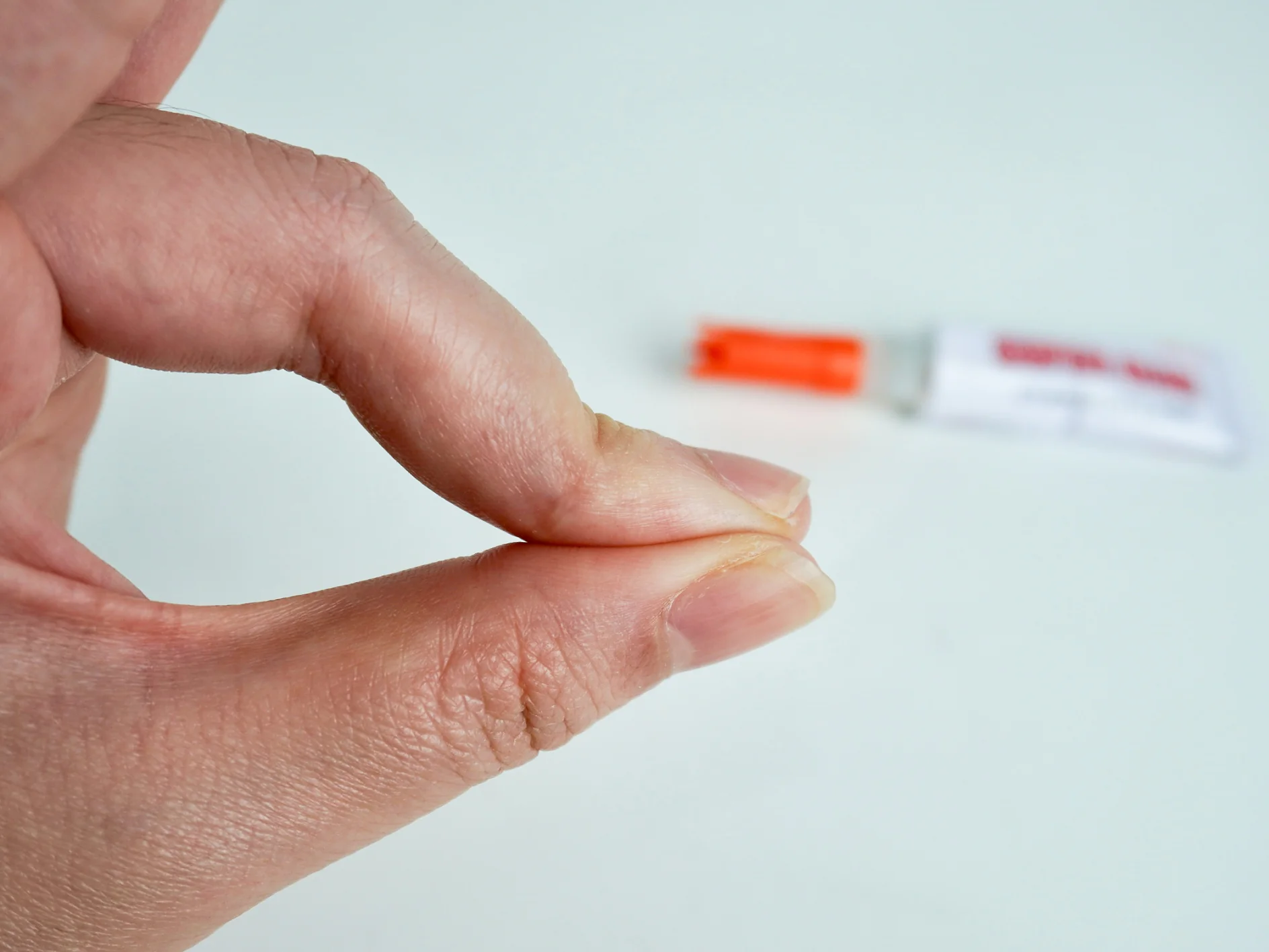 Eine Hand mit zusammengedrückten Fingern, als ob sie etwas Kleines drücken oder halten würde. Im Hintergrund, leicht unscharf, liegt eine Tube mit orangefarbener Kappe auf einer weißen Fläche neben einer Rolle Tesaband. (This text has been generated by AI)