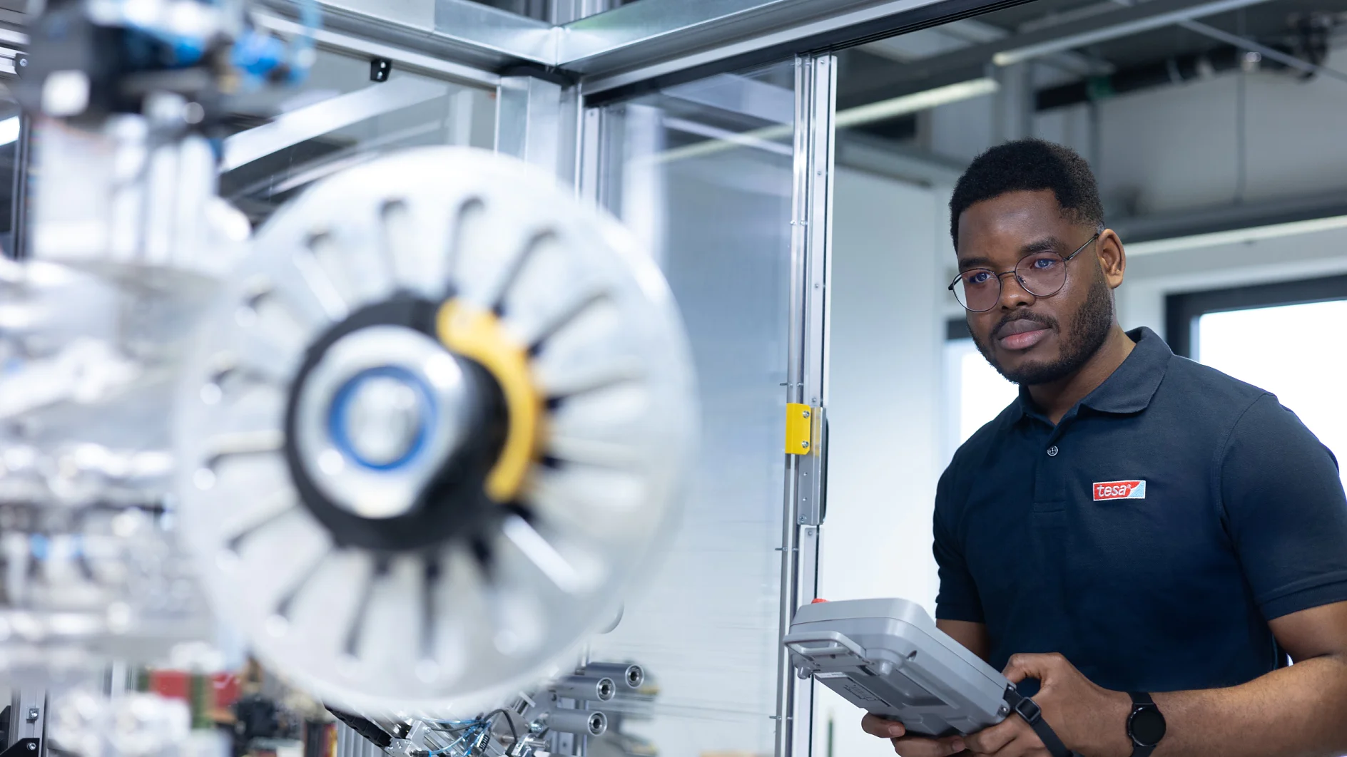 Eine Person in einem marineblauen Poloshirt und einem Tablet-PC steht neben Industrieanlagen in einer modernen Umgebung. Auf den verschiedenen Komponenten des großen mechanischen Rads im Vordergrund ist deutlich tesa-Klebeband zu sehen, während im Hintergrund Metallstrukturen und Glasplatten zu sehen sind. (This text has been generated by AI)
