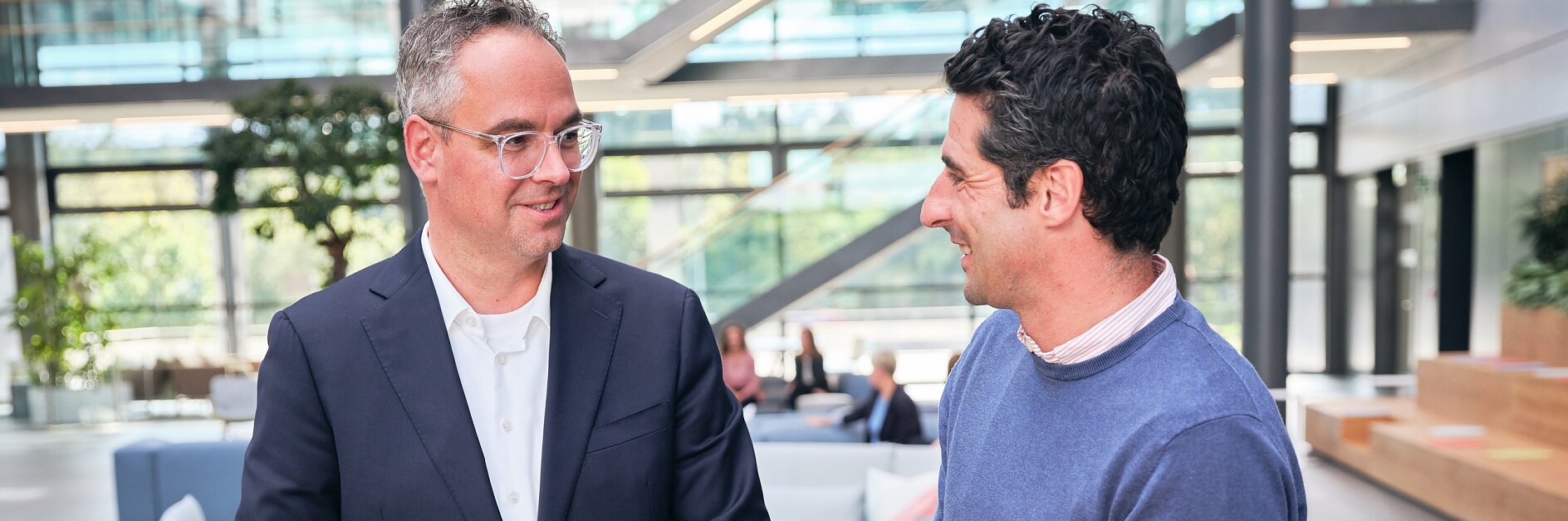 Zwei Männer stehen und unterhalten sich an einem Holztisch mit einem Laptop in einer Bürolobby. Einer der Männer trägt einen Anzug, der andere einen blauen Pullover. Ihre Unterhaltung scheint spannend zu sein. Im Hintergrund sind große Fenster und Sitzbereiche zu sehen. (This text has been generated by AI)